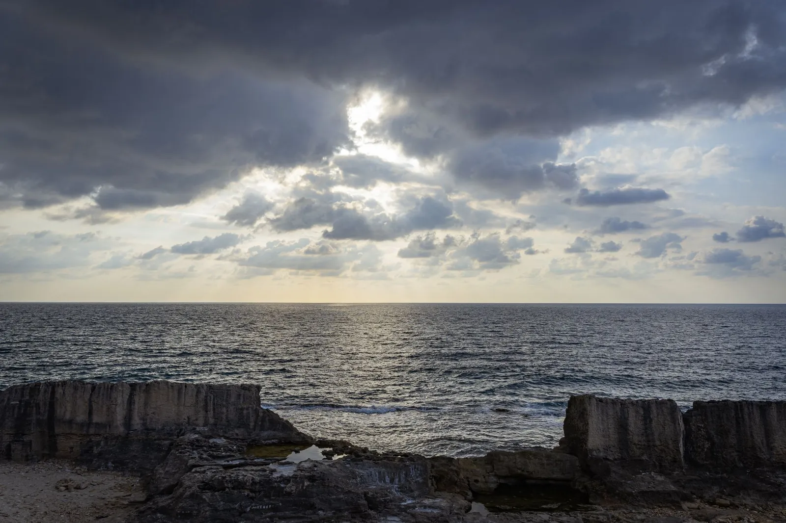 A wide view of a calm sea under a dramatic cloudy sky with sunlight breaking through, seen over a rocky coastline with cut stone blocks