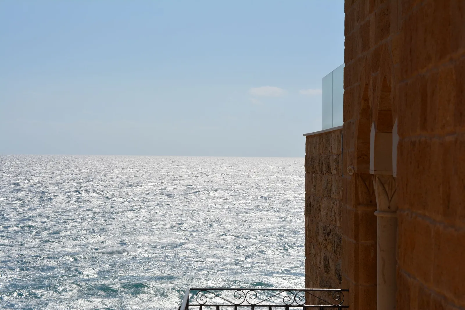 A sandstone building wall with pointed arches and a glass-panelled terrace edge beside a sparkling sea that reaches a clear horizon under a pale blue sky