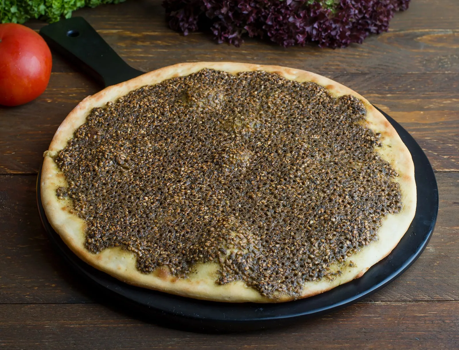 A round flatbread with a dark herb and sesame topping on a black round board with a handle
