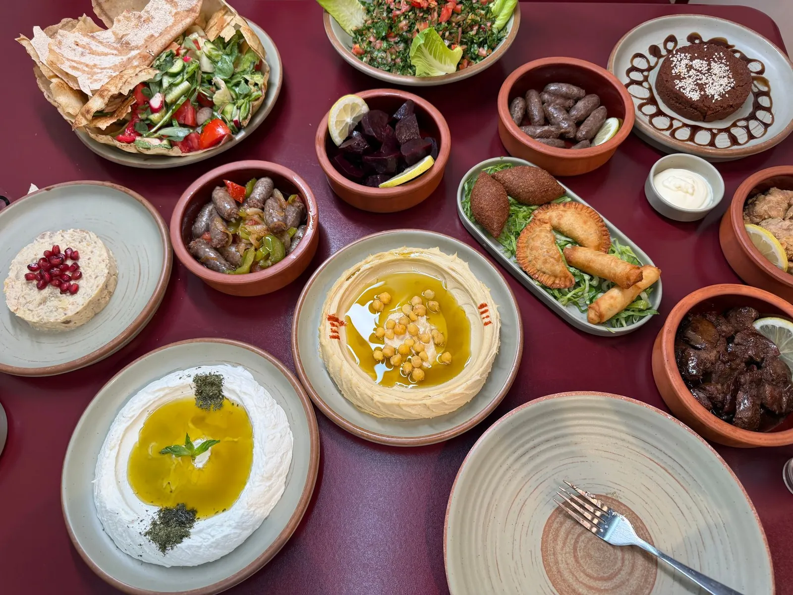 Top-down view of a dark red table covered with a mezze spread including hummus, labneh, tabbouleh, fattoush, grilled sausages, fried kibbeh and pastries in terracotta bowls and sage-green plates.