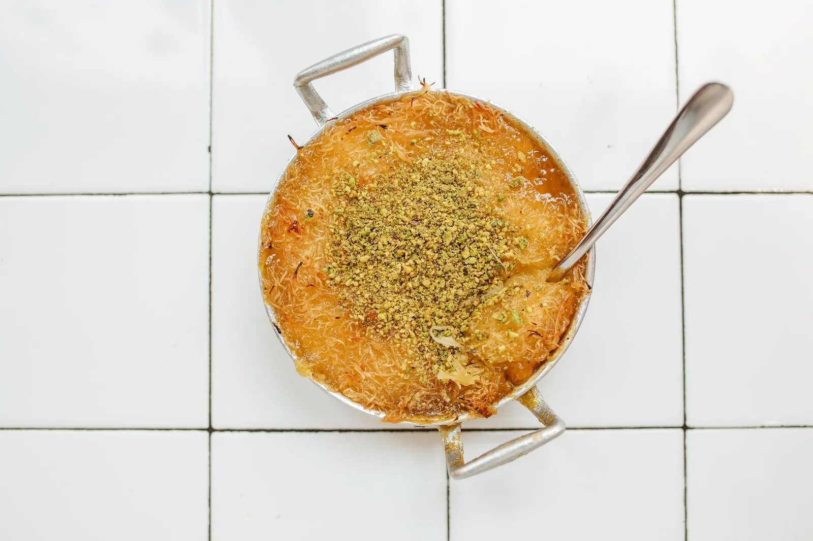 A small two-handled pan of golden knefe topped with crushed pistachios, with a spoon resting in it