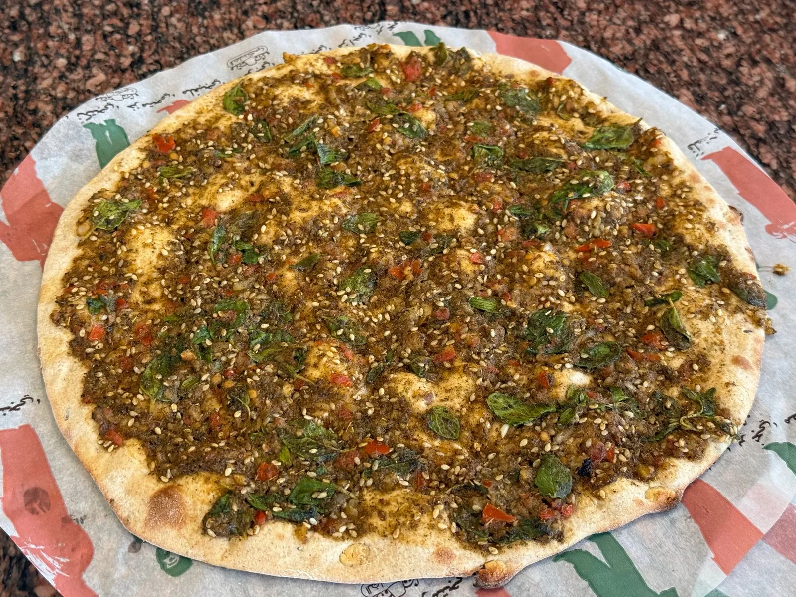 A round zaatar manakish with sesame seeds, green leaves and small pieces of red pepper on printed paper