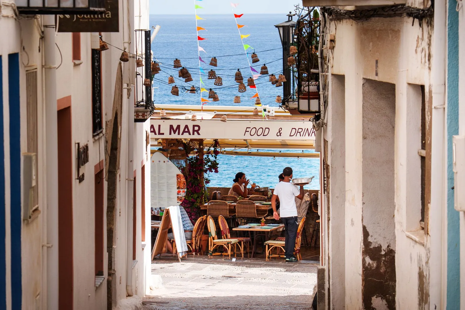 A whitewashed alley opening onto a seaside terrace with wicker chairs, hanging wicker lamps, diners at tables, colorful bunting and the blue sea behind
